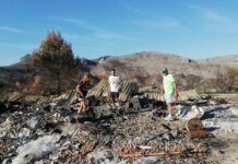 Drei Personen stehen auf einem abgebrannten Grundstück vor verbrannten Bergen.