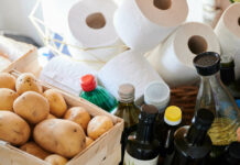 In einem Vorratszimmer stehen unterschiedliche Flaschen Öl, Essig, Toilettenpapier und ein Korb mit Kartoffeln.