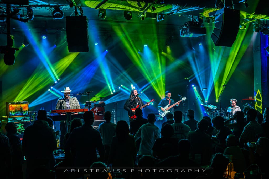 A wide-angle, medium shot captured from the back of a concert venue shows the band Pink Talking Fish performing on stage at The Hamilton Live. Four musicians are visible under a dramatic light show of crisscrossing yellow and blue beams that cut through the dark, hazy atmosphere. The band consists of: A keyboardist on the left wearing a fedora. A guitarist in the center singing into a microphone. A bassist to the right. A drummer in the far-right background. The silhouettes of a dense crowd are visible in the foreground, their backs to the camera as they watch the performance. The bottom of the image features a white watermark that reads "ARI STRAUSS PHOTOGRAPHY."