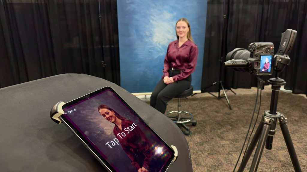 A headshot subject seated on a stool against a blue backdrop with a camera on a tripod and tablet preview
