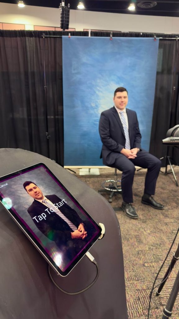 A man in a suit photographed on a stool against a blue backdrop, with his headshot previewed on a tablet