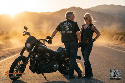AI photo experience: a guest couple styled as bikers in Rabble Rouser MC cuts stand with a Harley on an open desert road at sunset. Branded for Hideaway.