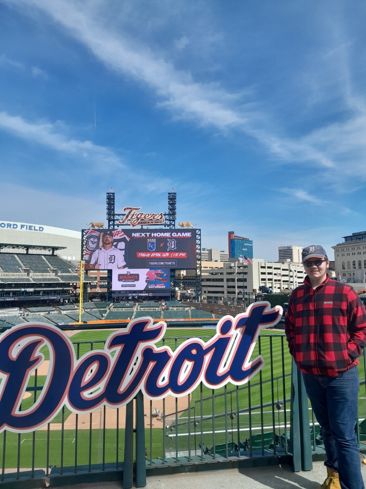 Photo of a the field baseball stadium , facing the scoreboard, in the background, with the Detroit logo at the bottom, and a 15-year-old young man wearing blue jeans and a buffalo plaid jacket with glasses and a ball cap in the foreground.