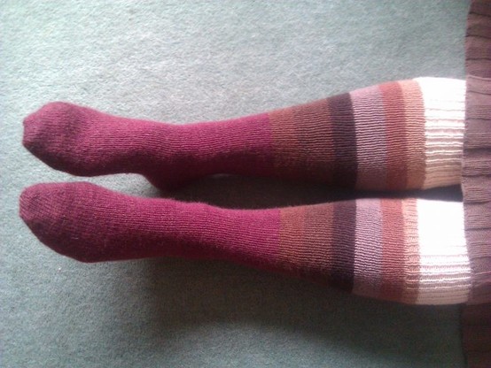 A pair of lower legs wearing hand knitted socks. The socks progress from foot to just below the knee in a sequence of colour blocks: brownish red, browns, cream. The blocks are not quite lined up on the two socks.